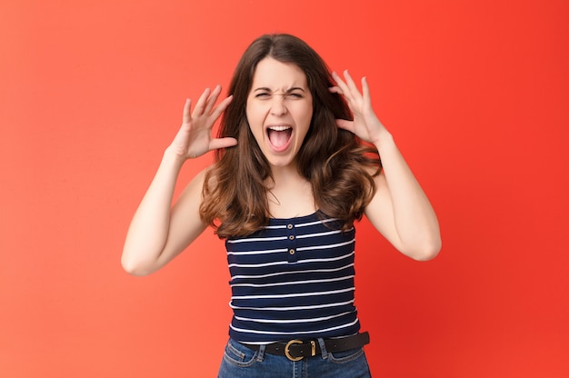 Young pretty woman screaming in panic or anger, shocked, terrified or furious, with hands next to head over red wall