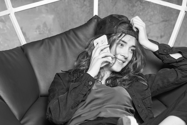 Young pretty woman or sexy smiling girl with long hair in black cap and sneakers sitting in gesometric chair with mobile or cell phone on grey background