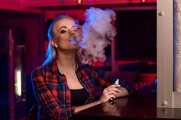 Young pretty woman in a shirt in a cage smoke an electronic cigarette at the vape bar