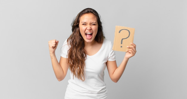 Young pretty woman shouting aggressively with an angry expression and holding a question mark sign