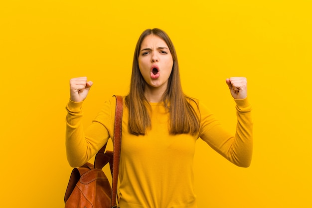 Young pretty woman shouting aggressively with an angry expression or with fists clenched celebrating success against orange 