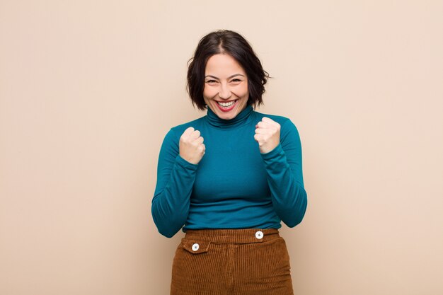 Young pretty woman shouting triumphantly, laughing and feeling happy and excited while celebrating success against beige wall