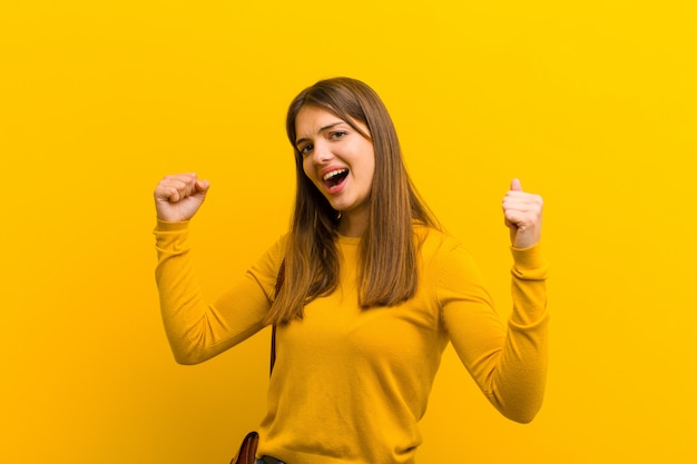 Young pretty woman shouting triumphantly, looking like excited, happy and surprised winner, celebrating against orange wall