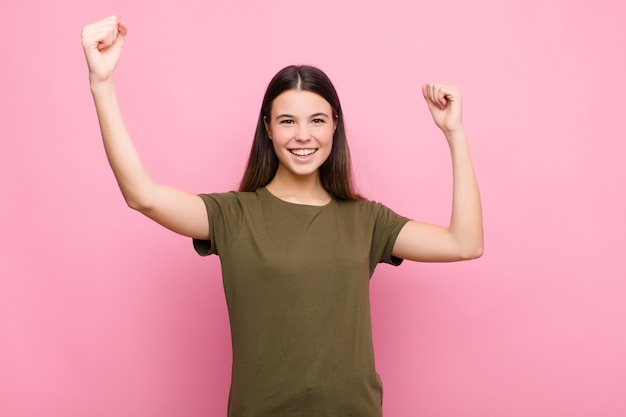 Young pretty woman shouting triumphantly, looking like excited, happy and surprised winner, celebrating against pink wall