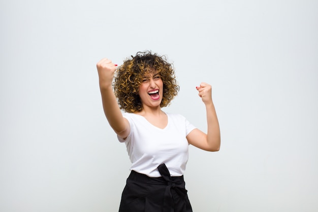 Young pretty woman shouting triumphantly, looking like excited, happy and surprised winner, celebrating against white wall