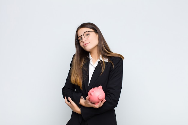 Young pretty woman shrugging, feeling confused and uncertain, doubting with arms crossed and puzzled look with a piggy bank