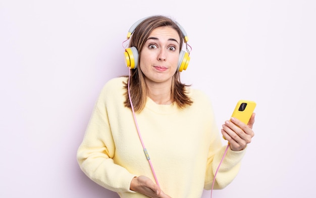 Young pretty woman shrugging, feeling confused and uncertain. headphones and phone concept