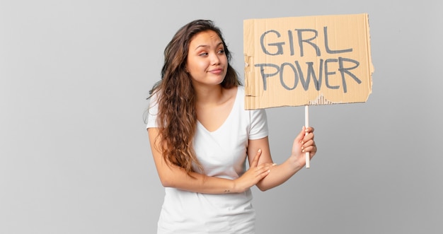 Young pretty woman shrugging, feeling confused and uncertain and holding a girl power banner