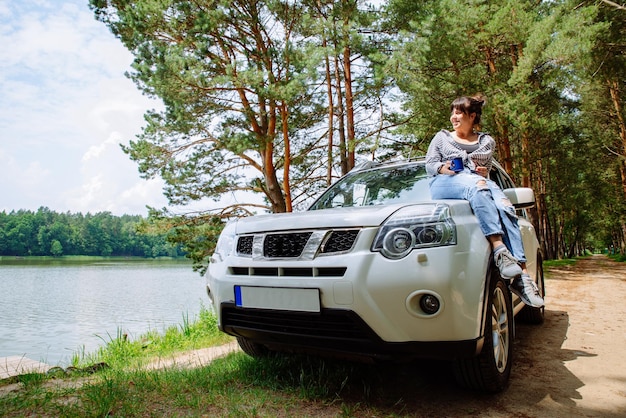 Young pretty woman sitting on car hood drinking coffee lake with forest on background