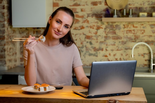 La giovane donna graziosa che si siede alla sua cucina mangia il sushi al tavolo a casa lavorando con un computer portatile