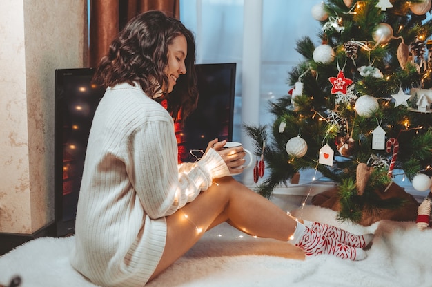 Young pretty woman sitting near christmas tree drinking cup of coffee copy space