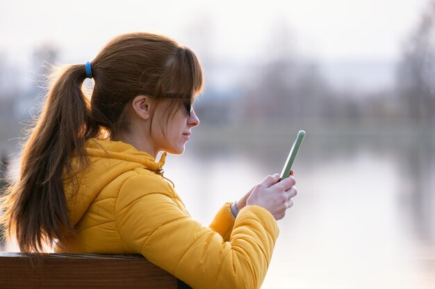 夕方に屋外で彼女の携帯電話を閲覧している公園のベンチに座っている若いきれいな女性。