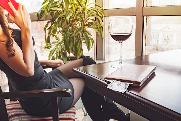 Молодая красивая женщина сидит в ресторане, пьет бокал вина, смотрит в панорамное окно с прекрасным видом на город и разговаривает по телефону. 100 долларов в папке для счета на столе.