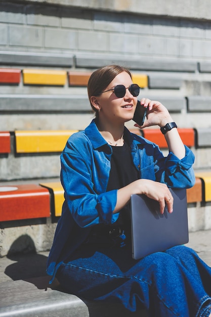Молодая красивая женщина сидит с ноутбуком на современной парковой улице с помощью смартфона на удаленной ИТ-работе в летнем стиле, небрежно улыбаясь в очках