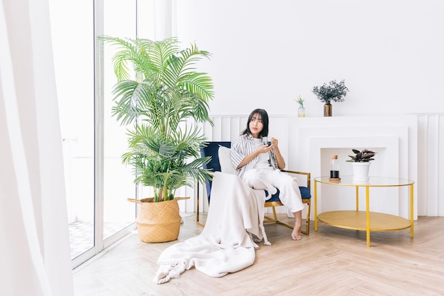 Young pretty woman smile and sits in the chair of the modern\
living area with a green plant relaxing drinking a cup of hot\
coffee and enjoying a quiet time and indoor environment at cozy\
home