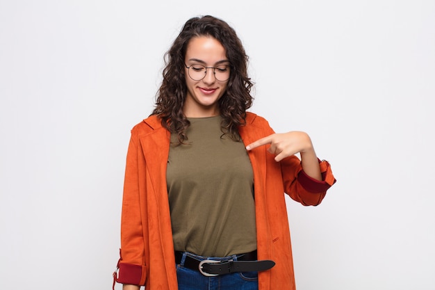 Foto giovane donna graziosa che sorride allegramente e casualmente, guardando verso il basso e indicando il petto contro il muro bianco