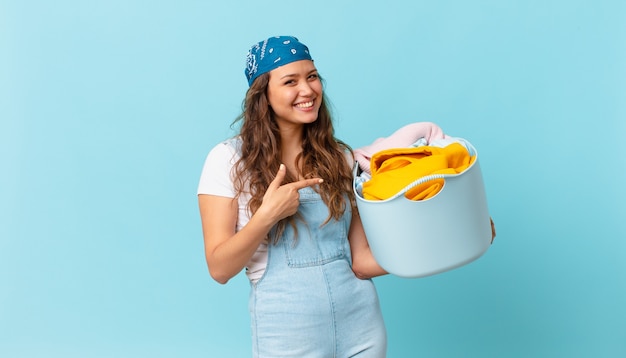 Young pretty woman smiling cheerfully, feeling happy and pointing to the side and holding a wash clothes basket