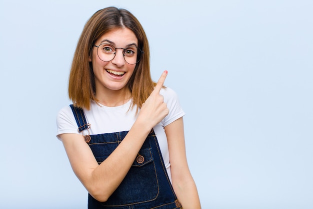 Young pretty woman smiling cheerfully, feeling happy and pointing to the side and upwards, showing object in copy space on blue