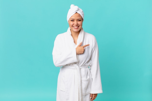 Young pretty woman smiling cheerfully, feeling happy and pointing to the side and wearing bath robe after shower