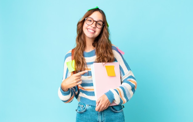 Giovane bella donna che sorride allegramente, si sente felice e indica il lato con una borsa e tiene in mano dei libri