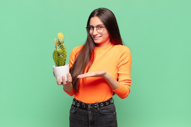 Young pretty woman smiling cheerfully, feeling happy and showing a concept in copy space with palm of hand. cactus concept