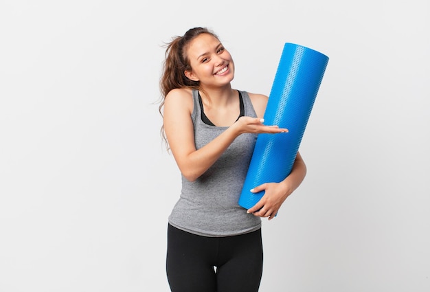 Young pretty woman smiling cheerfully, feeling happy and showing a concept and holding a yoga mat