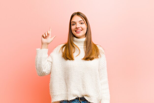 Young pretty woman smiling cheerfully and happily, pointing upwards with one hand to copy space against pink background