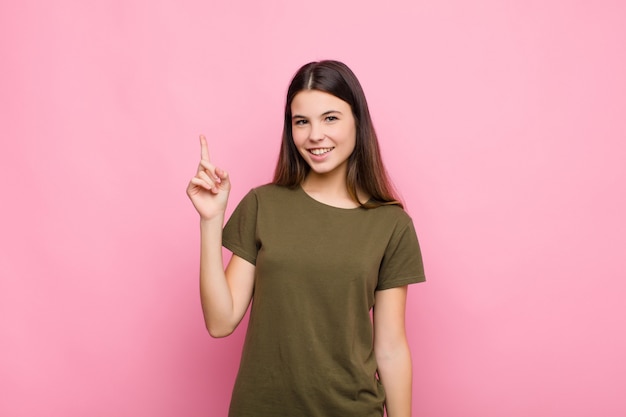 Young pretty woman smiling cheerfully and happily, pointing upwards with one hand to copy space against pink wall