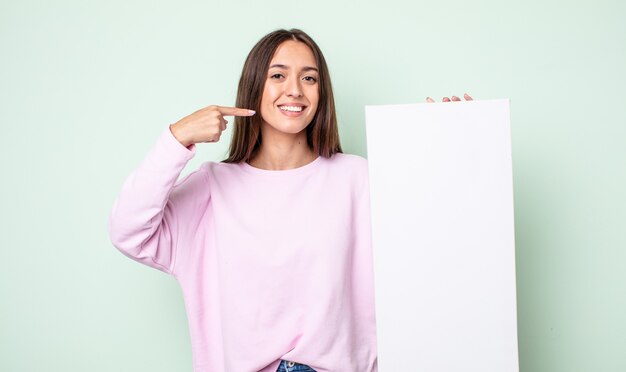 Young pretty woman smiling confidently pointing to own broad smile. empty canvas concept