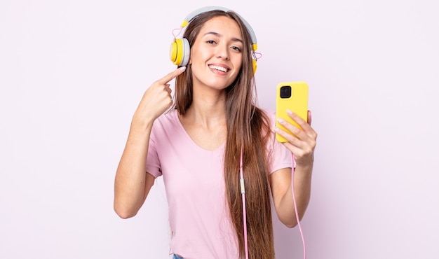 Young pretty woman smiling confidently pointing to own broad smile. headphones and smartphone