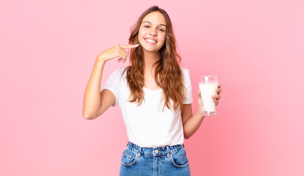 自信を持って笑顔の若いきれいな女性は、自分の広い笑顔を指して、ミルクのグラスを持っています
