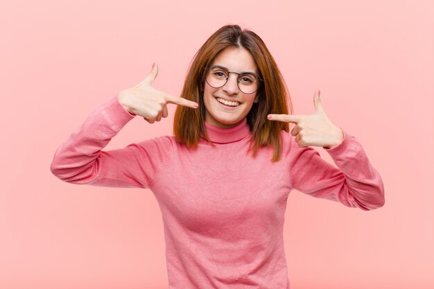 Young pretty woman smiling confidently pointing to own broad smile, positive, relaxed, satisfied attitude against pink wall