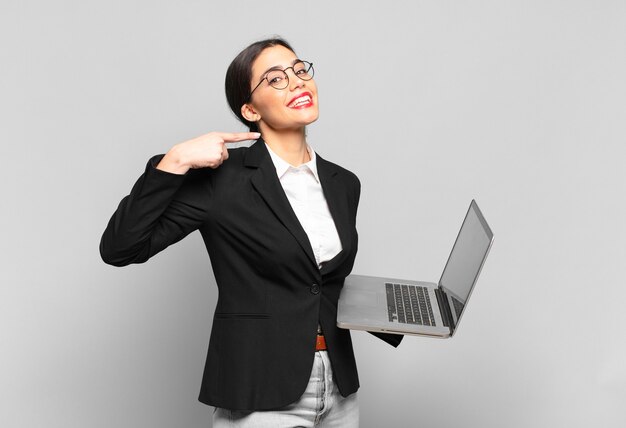 Young pretty woman smiling confidently pointing to own broad smile, positive, relaxed, satisfied attitude. laptop concept