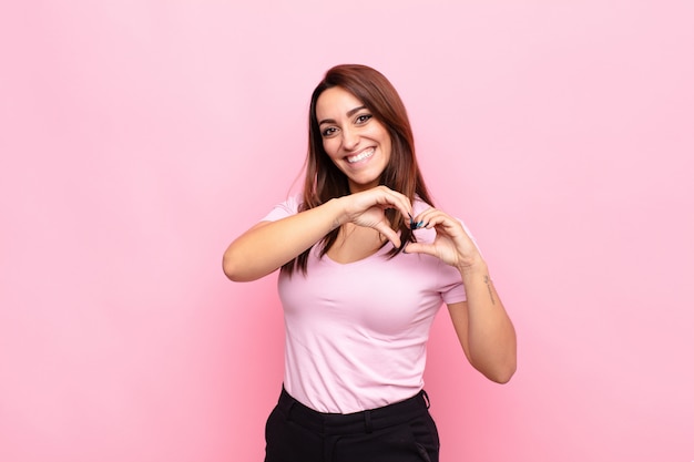 Young pretty woman smiling and feeling happy, cute, romantic and in love, making heart shape with both hands against pink wall