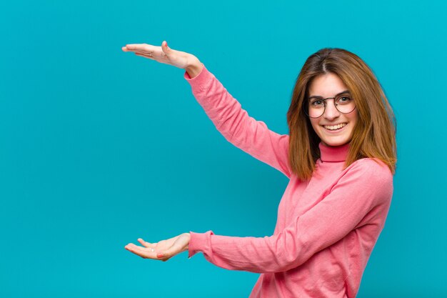 Young pretty woman smiling, feeling happy, positive and satisfied, holding or showing object or concept on copy space against blue background