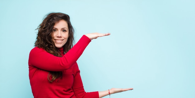 若いきれいな女性の笑みを浮かべて、幸せ、肯定的な満足を感じて、保持または青い壁に対してコピースペース上のオブジェクトまたは概念を示す