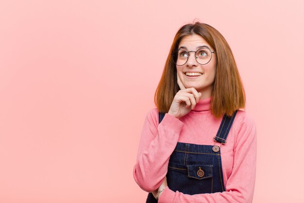 Giovane donna graziosa che sorride felicemente e sognare ad occhi aperti o dubitare, guardando al lato sul rosa