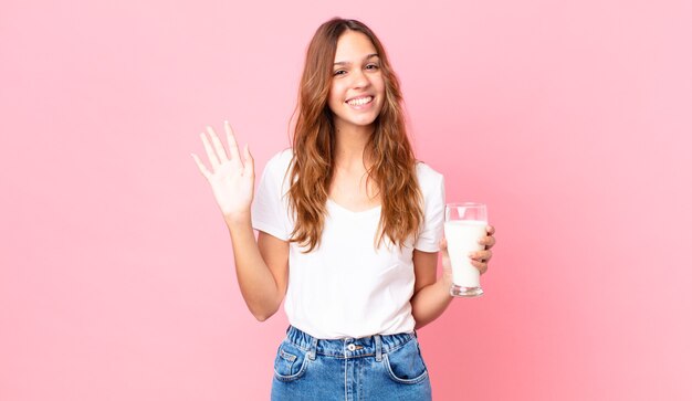 Young pretty woman smiling happily, waving hand, welcoming and greeting you and holding a glass of milk