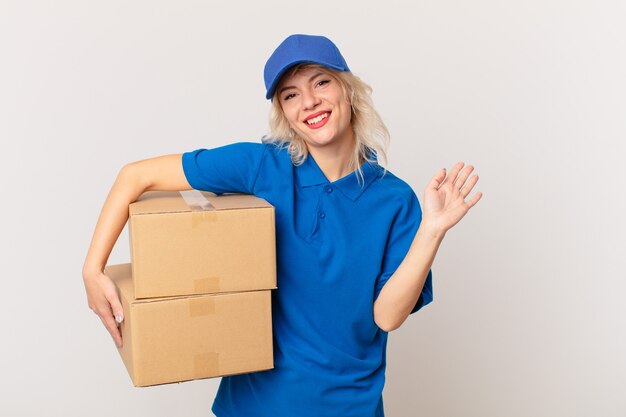 Photo young pretty woman smiling happily, waving hand, welcoming and greeting you. package delivering concept