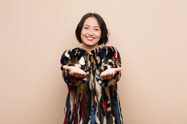 Young pretty woman smiling happily with friendly, confident, positive look, offering and showing an object or concept against beige wall