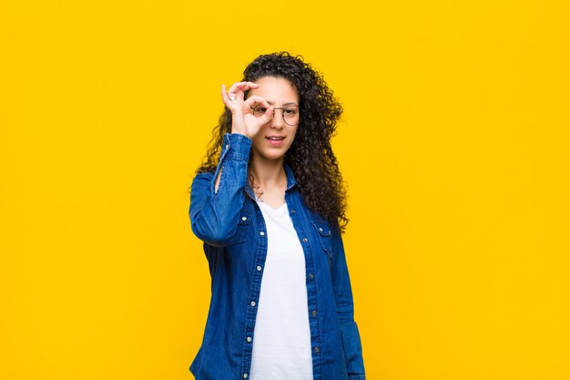 Young pretty woman smiling happily with funny face, joking and looking through peephole, spying on secrets against orange wall