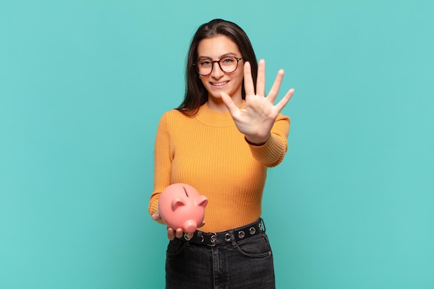 Young pretty woman smiling and looking friendly, showing number five or fifth with hand forward, counting down. piggy bank concept