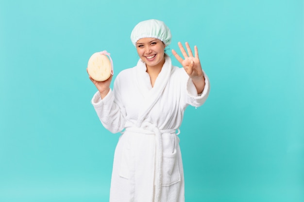 Young pretty woman smiling and looking friendly, showing number four and wearing bath robe after shower