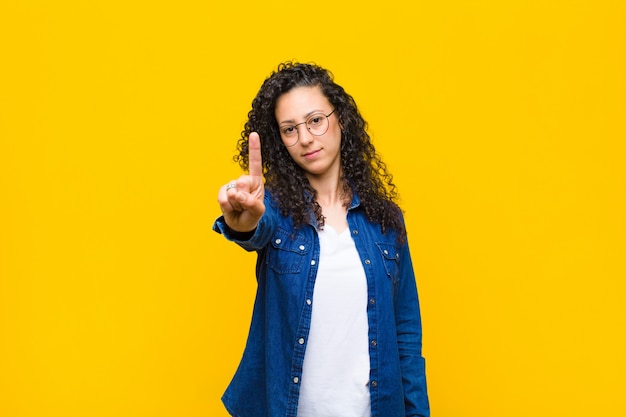 Foto giovane donna graziosa che sorride e che sembra amichevole, mostrando numero uno o primo con la mano in avanti, conto alla rovescia contro la parete arancione