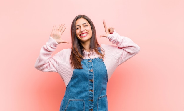 Young  pretty woman smiling and looking friendly, showing number seven or seventh with hand forward, counting down over pink wall.