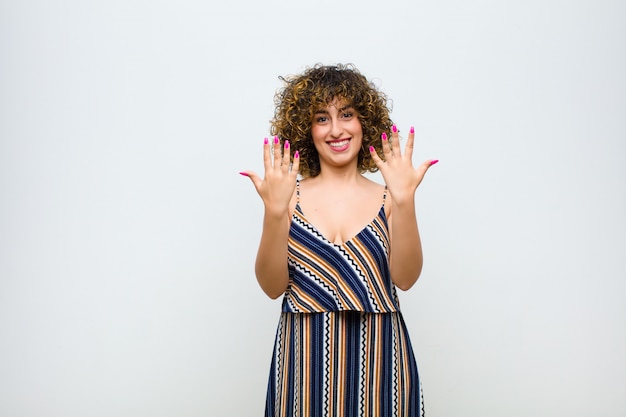 Young pretty woman smiling and looking friendly, showing number ten or tenth with hand forward, counting down against white wall