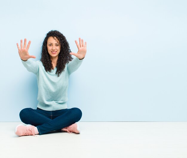 Young pretty woman smiling and looking friendly, showing number ten or tenth with hand forward, counting down sitting on floor