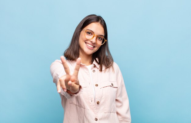 Young pretty woman smiling and looking happy, carefree and positive, gesturing victory or peace