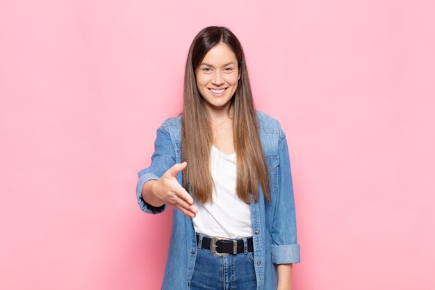 Giovane donna graziosa che sorride, sembra felice, sicura e amichevole, offrendo una stretta di mano per concludere un affare, collaborando