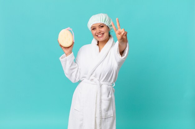 Young pretty woman smiling and looking happy, gesturing victory or peace and wearing bath robe after shower